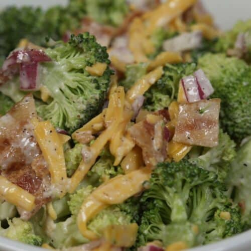 Close-up of an Amish broccoli salad with shredded cheddar cheese, red onion, bacon pieces, and a creamy dressing in a white bowl.