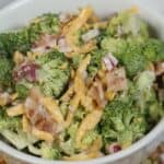 A bowl of Amish broccoli salad with shredded cheddar cheese, chopped red onions, and bacon pieces, on a patterned surface.
