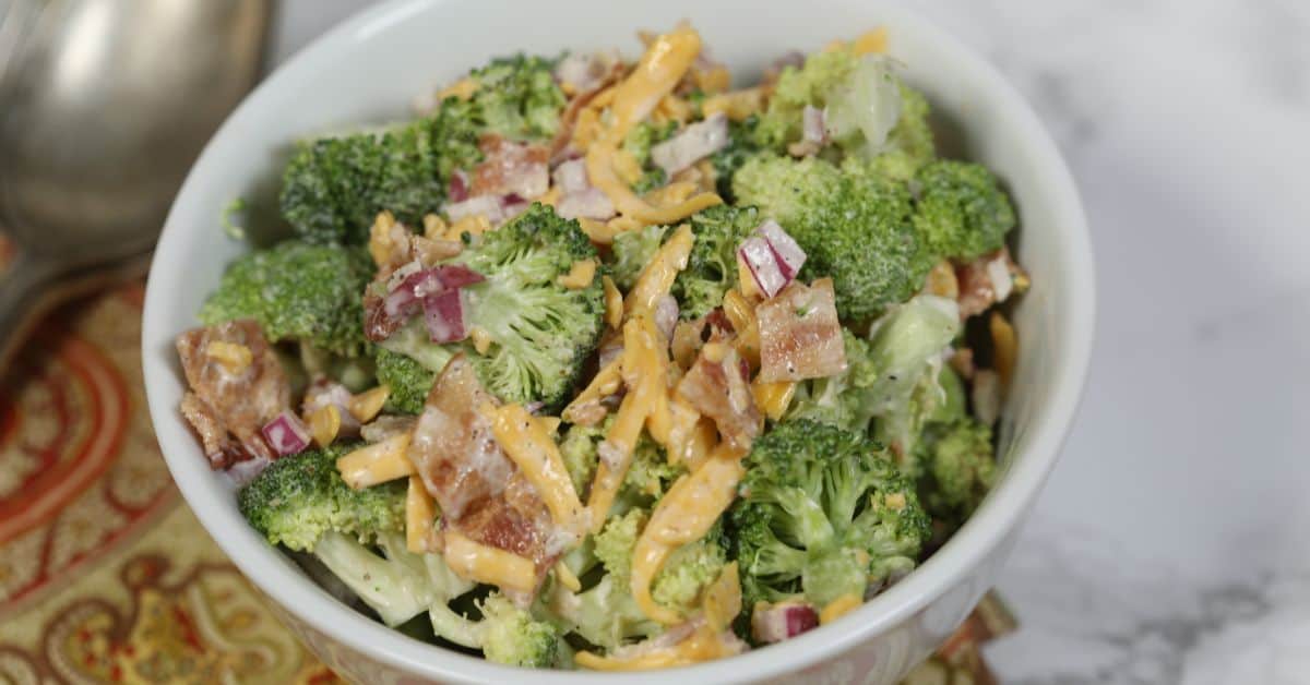 A bowl of Amish broccoli salad with shredded cheddar cheese, chopped red onions, and bacon pieces, on a patterned surface.