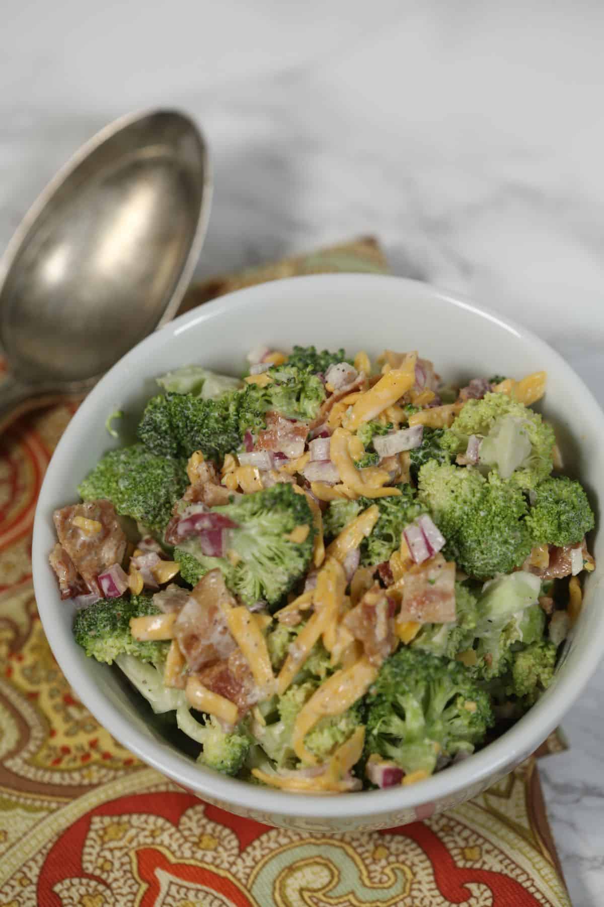 A white bowl of Amish broccoli salad with bacon, shredded cheese, and red onion sits on a patterned cloth next to a metal spoon.