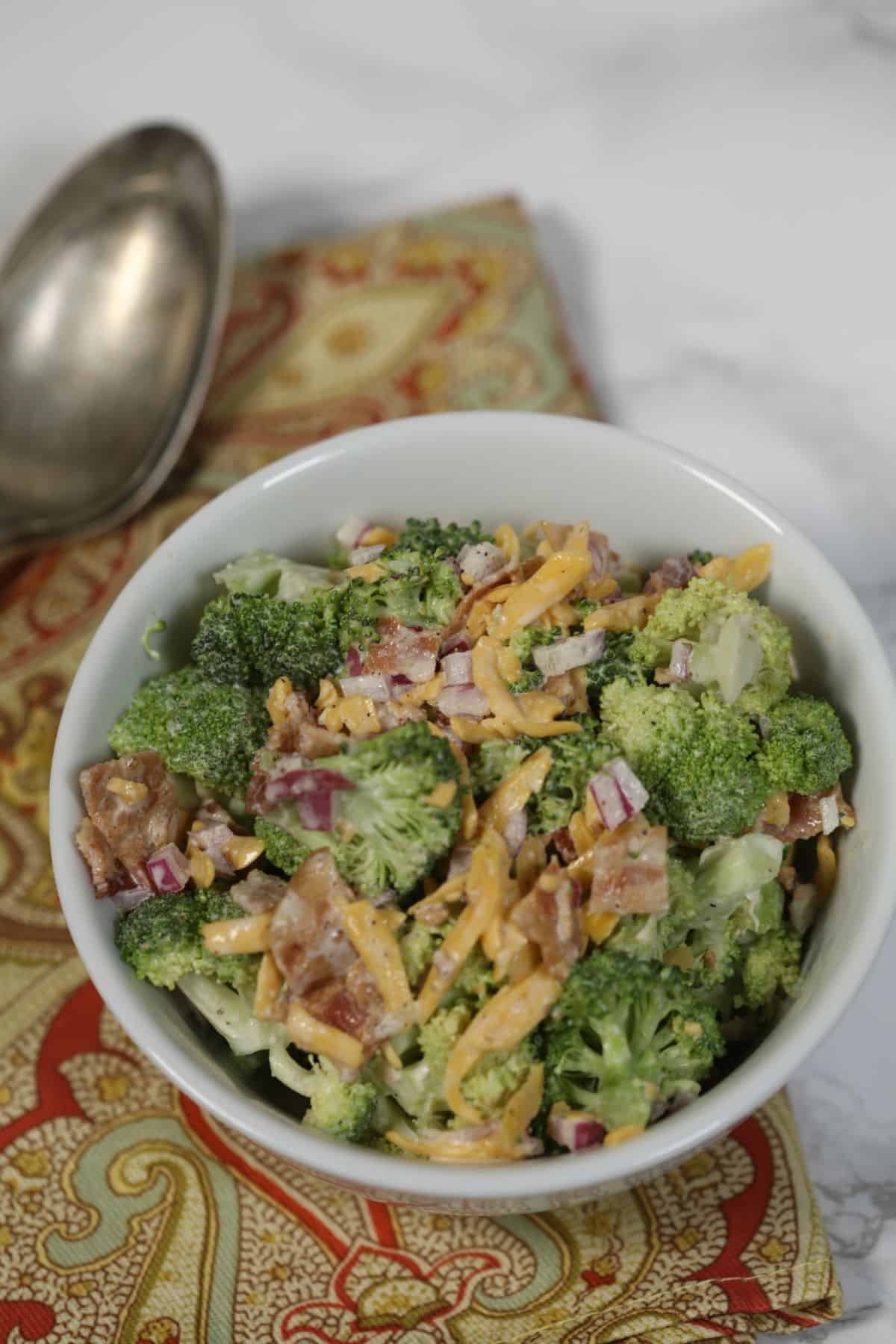 A bowl of Amish broccoli salad with shredded cheddar cheese, bacon, and red onion, placed on a patterned napkin with a metal spoon beside it.