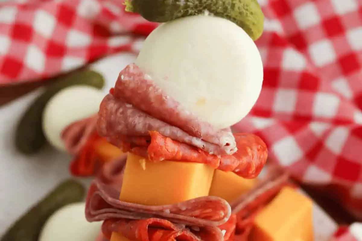 A close-up of a kabob stack with cheddar cheese cubes, slices of salami and pepperoni, a mozzarella ball, and a small pickle on top, with a red and white checkered cloth in the background—perfect for easy kabob recipes.