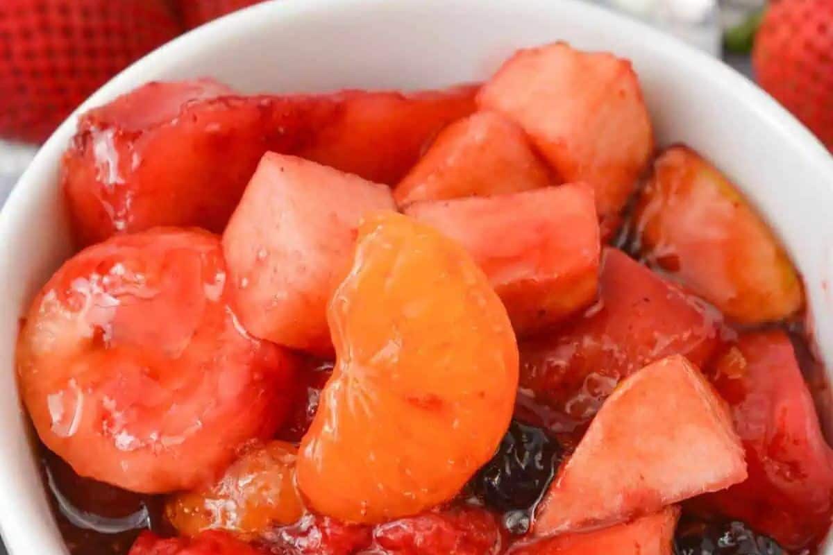 A close-up of a white bowl filled with fruit salad, featuring pieces of banana, orange, watermelon, and berries in a red syrup—one of the most mouthwatering salads perfect for summer.