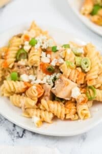 Plate of rotini chicken wing pasta salad with pieces of chicken, topped with crumbled cheese, chopped green onions, and an orange sauce.