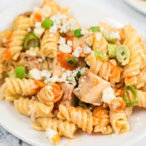 Plate of rotini chicken wing pasta salad with pieces of chicken, topped with crumbled cheese, chopped green onions, and an orange sauce.