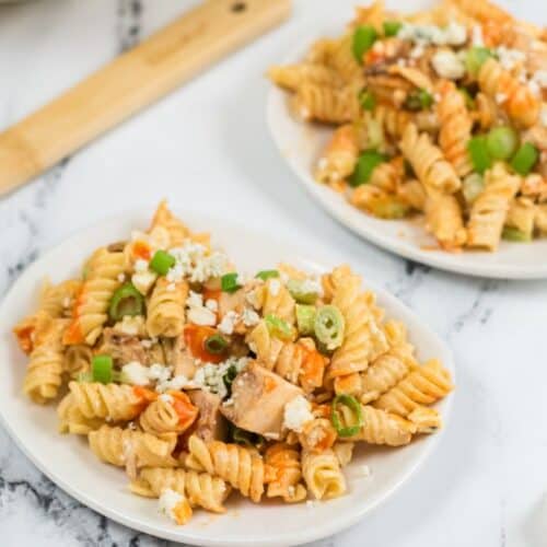Two white plates with buffalo chicken pasta salad—featuring rotini pasta, diced chicken, green onions, and crumbled cheese—rest on a marble surface next to a wooden spatula and a red-striped napkin.