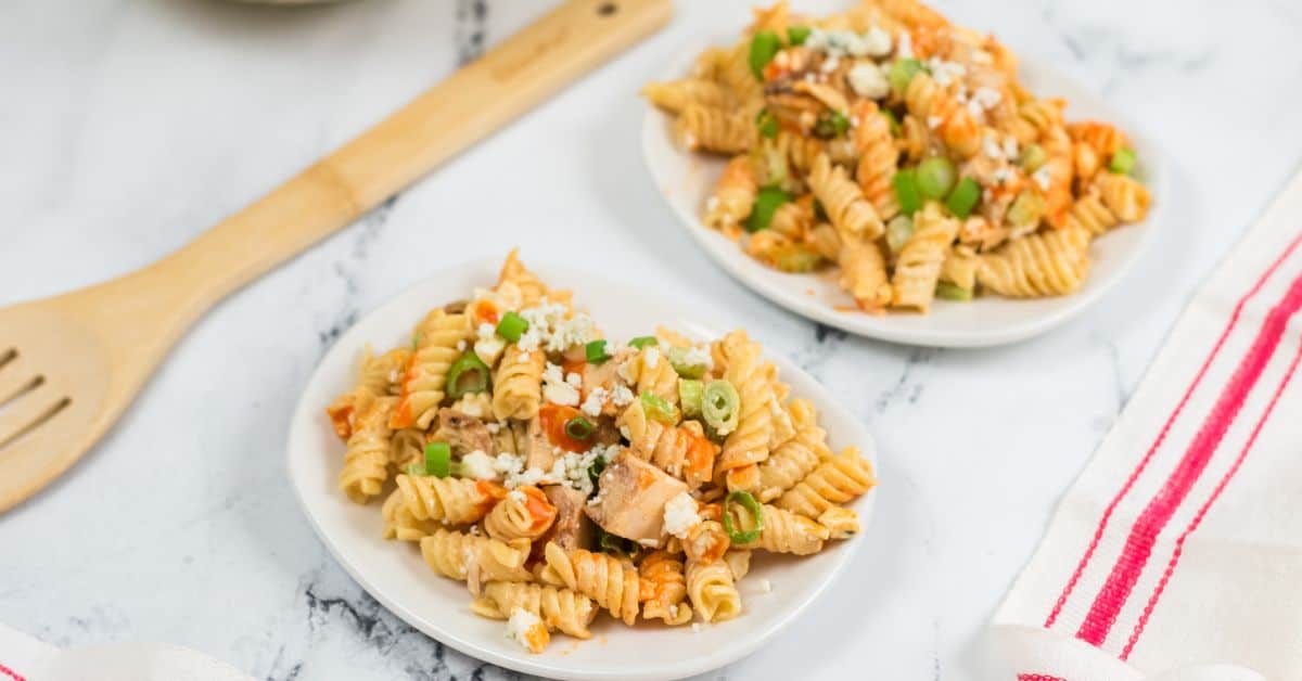 Two white plates with buffalo chicken pasta salad—featuring rotini pasta, diced chicken, green onions, and crumbled cheese—rest on a marble surface next to a wooden spatula and a red-striped napkin.