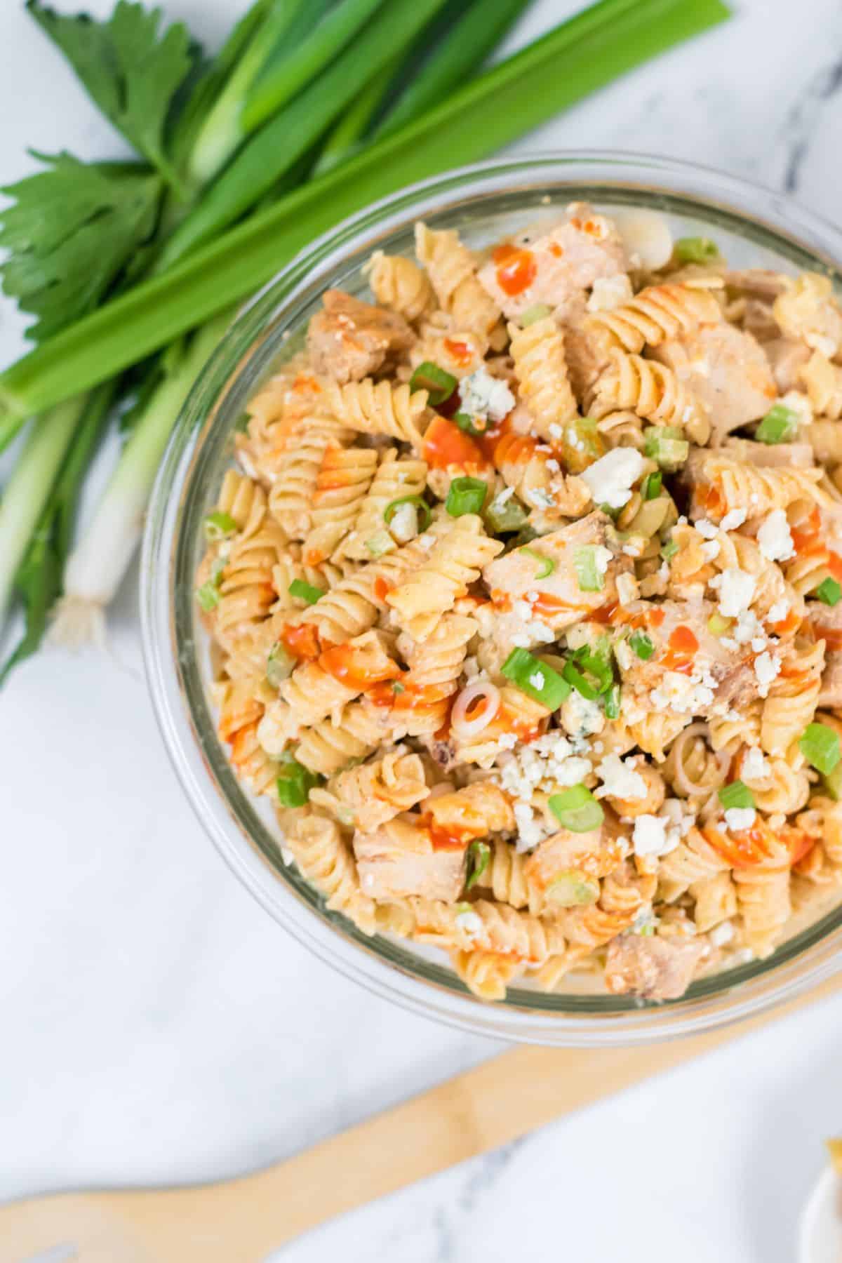 A bowl of buffalo chicken pasta salad with rotini, green onions, crumbled cheese, and hot sauce sits next to fresh green onions and parsley on a white surface.