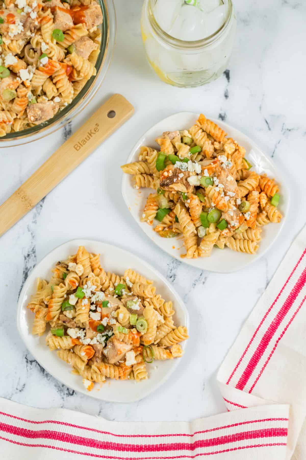 Two plates of buffalo chicken pasta salad with rotini, vegetables, and cheese are on a marble surface next to a bowl, a wooden spatula, a glass of water, and a red-striped towel.