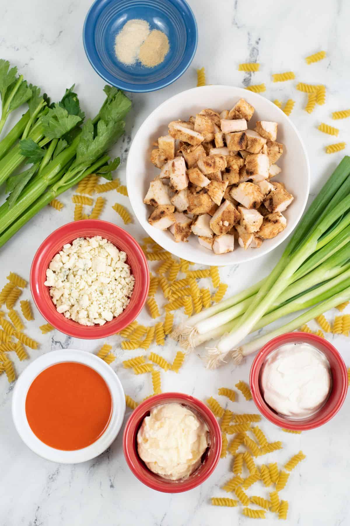 Ingredients for a buffalo chicken pasta salad: cooked chicken, blue cheese, celery, green onions, pasta, buffalo sauce, mayonnaise, sour cream, and seasonings arranged on a white marble surface create the perfect base for this flavorful buffalo chicken pasta salad.