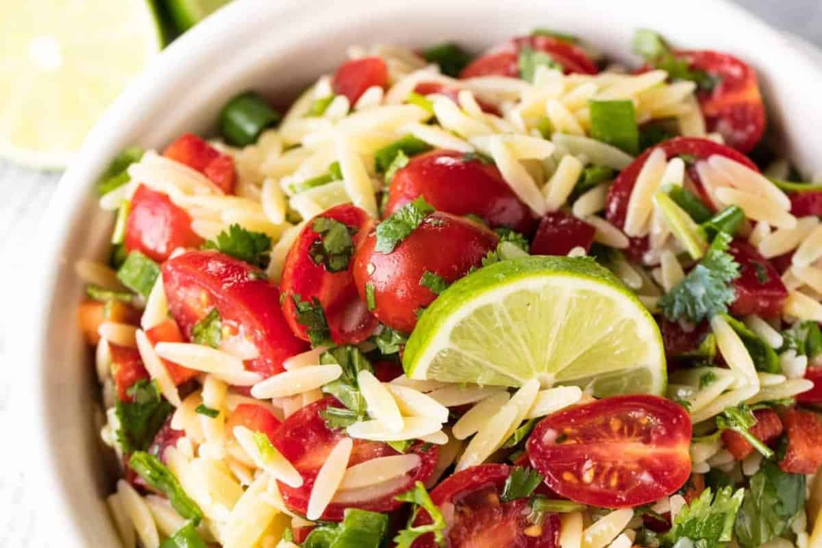 A bowl of orzo pasta salad with cherry tomatoes, chopped green herbs, and a slice of lime on top—one of the most refreshing summer salads for your collection.