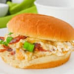 A sandwich on a white plate with a bun, filled with a mixture of creamy spread, bacon, and chopped green onions. A green cloth is in the background. This delectable Cracked Chicken Sandwich is sure to delight your taste buds.