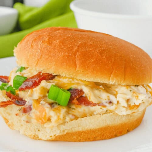 A sandwich on a white plate with a bun, filled with a mixture of creamy spread, bacon, and chopped green onions. A green cloth is in the background. This delectable Cracked Chicken Sandwich is sure to delight your taste buds.