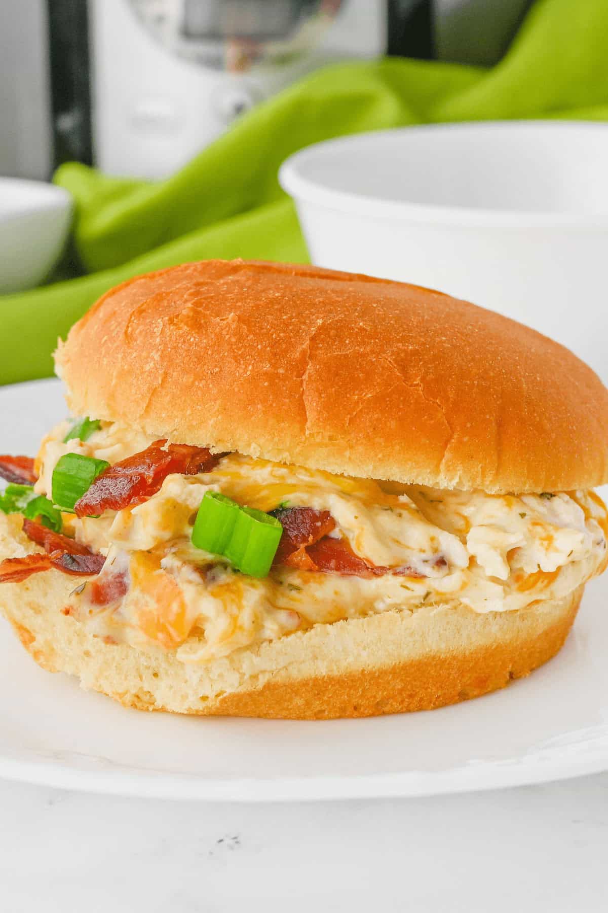 A sandwich on a white plate with a bun, filled with a mixture of creamy spread, bacon, and chopped green onions. A green cloth is in the background. This delectable Cracked Chicken Sandwich is sure to delight your taste buds.