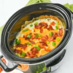 A slow cooker filled with a cheesy casserole topped with bacon bits and chopped green onions, reminiscent of the flavors in cracked chicken sandwiches. Shredded cheese and additional green onions are in small separate bowls beside the slow cooker.