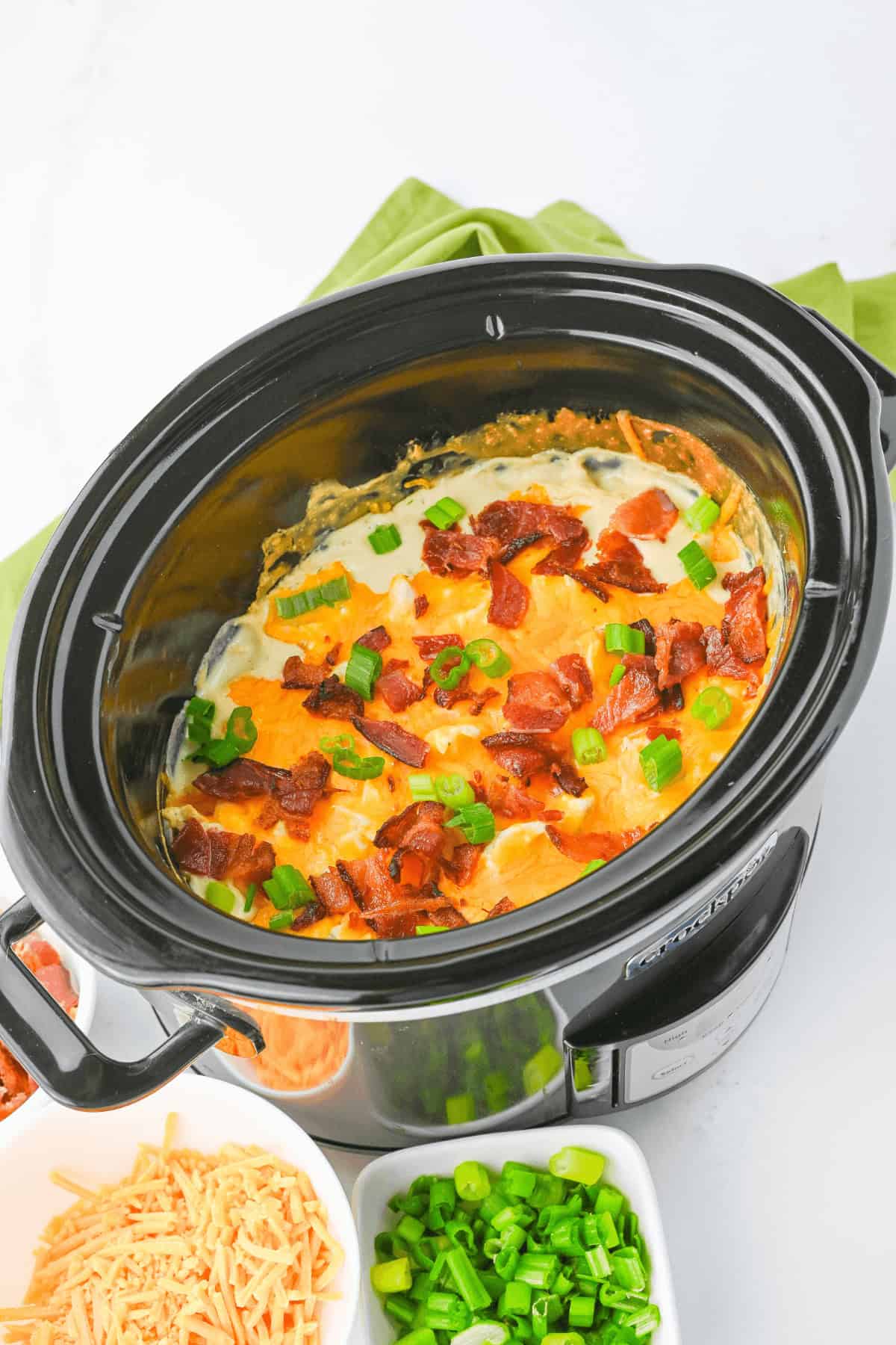 A slow cooker filled with a cheesy casserole topped with bacon bits and chopped green onions, reminiscent of the flavors in cracked chicken sandwiches. Shredded cheese and additional green onions are in small separate bowls beside the slow cooker.