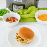 A pot with the meat sits on a white plate in front of a crockpot. Nearby are bowls with chopped bacon, shredded cheese, and green onions.
