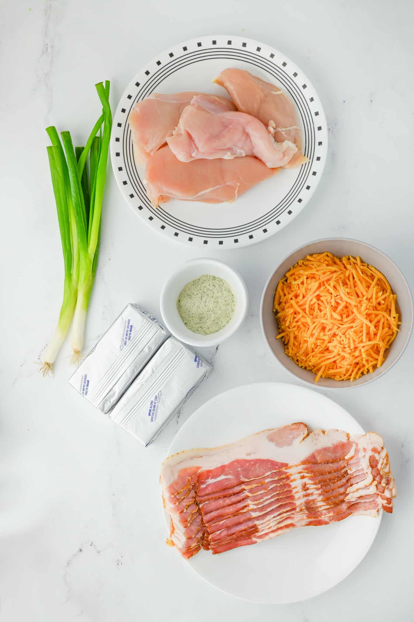 Ingredients on a counter: green onions, cream cheese blocks, chicken breasts on a plate, shredded cheese in a bowl, bacon slices on a plate, and a small dish of seasoning for the perfect Cracked Chicken Sandwiches.