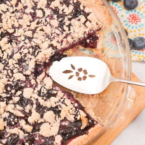 A baked Crumble Top Blueberry Pie with a missing slice, a pie server resting in the dish, and fresh blueberries scattered nearby on a patterned cloth.