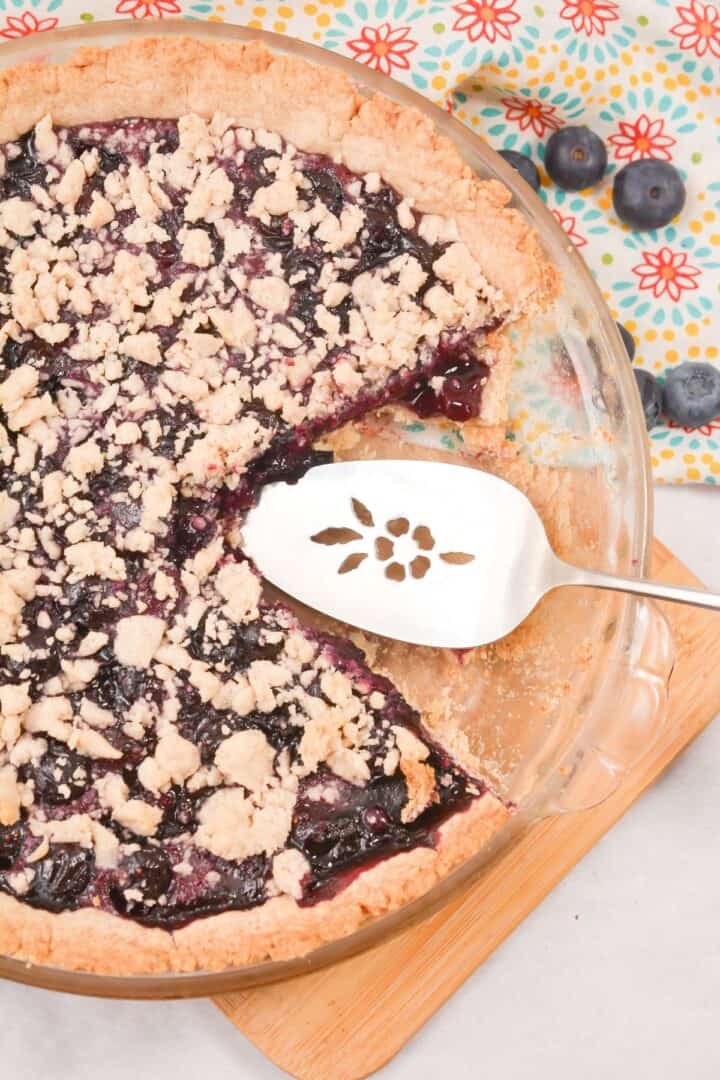 A baked Crumble Top Blueberry Pie with a missing slice, a pie server resting in the dish, and fresh blueberries scattered nearby on a patterned cloth.