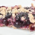 A slice of Crumble Top Blueberry Pie sits on a white plate, highlighting its flaky crust, vibrant blueberry filling, and deliciously crumbly topping.