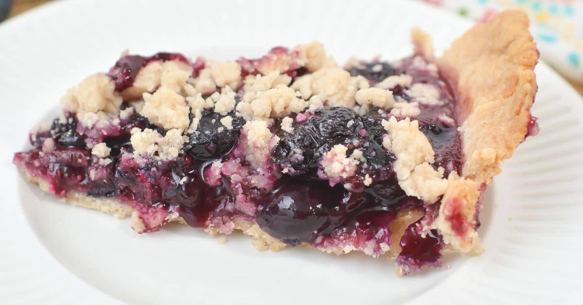 A slice of Crumble Top Blueberry Pie sits on a white plate, highlighting its flaky crust, vibrant blueberry filling, and deliciously crumbly topping.