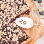 A Crumble Top Blueberry Pie with one slice missing sits in a glass pie dish, a pie server beside it on a wooden board. Fresh blueberries are scattered nearby, highlighting the fruity delight.