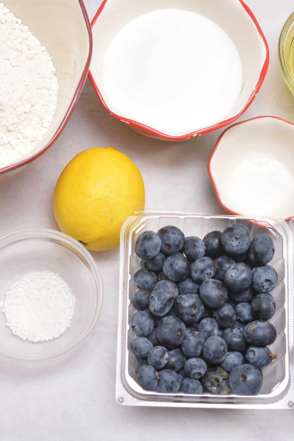 A plastic container of blueberries, a lemon, and bowls containing flour, sugar, salt, and other baking ingredients arranged on a white surface.