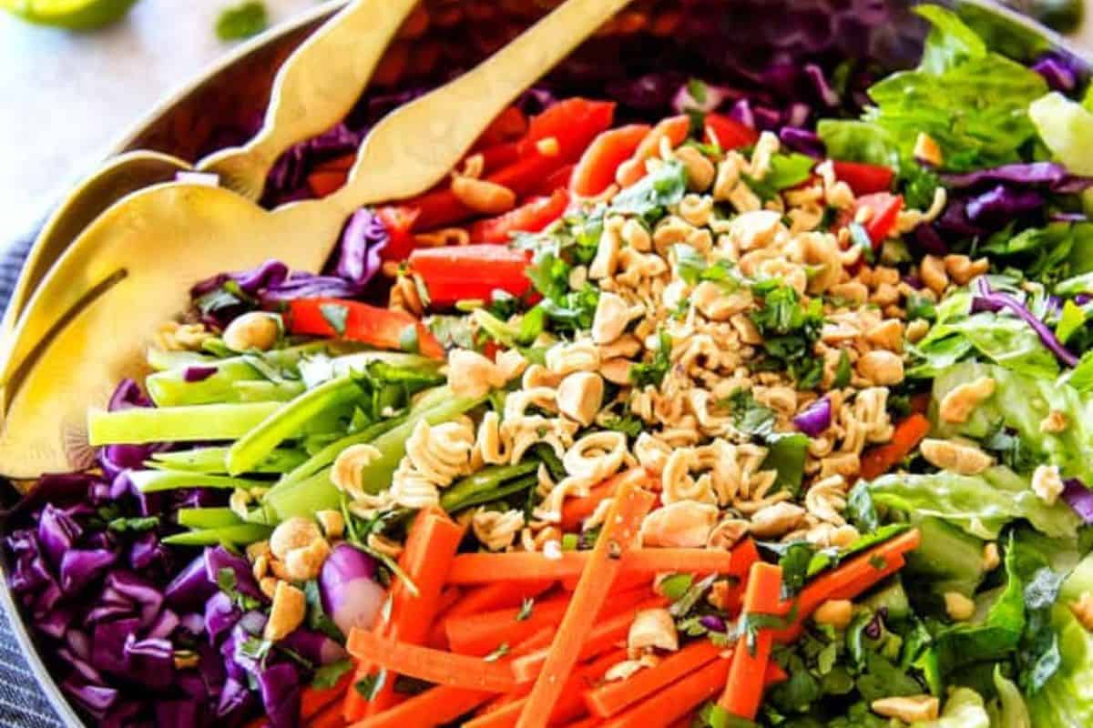 A colorful summer salad with chopped lettuce, red cabbage, carrots, cucumber, red bell pepper, peanuts, herbs, and gold salad servers—perfect for those who love mouthwatering salads.