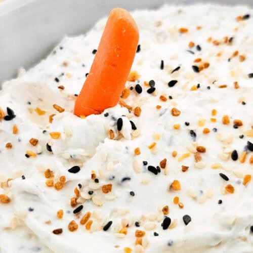 A baby carrot stands upright in a bowl of everything bagel dip, garnished with a mix of black and white sesame seeds. Blurred carrots are in the background.
