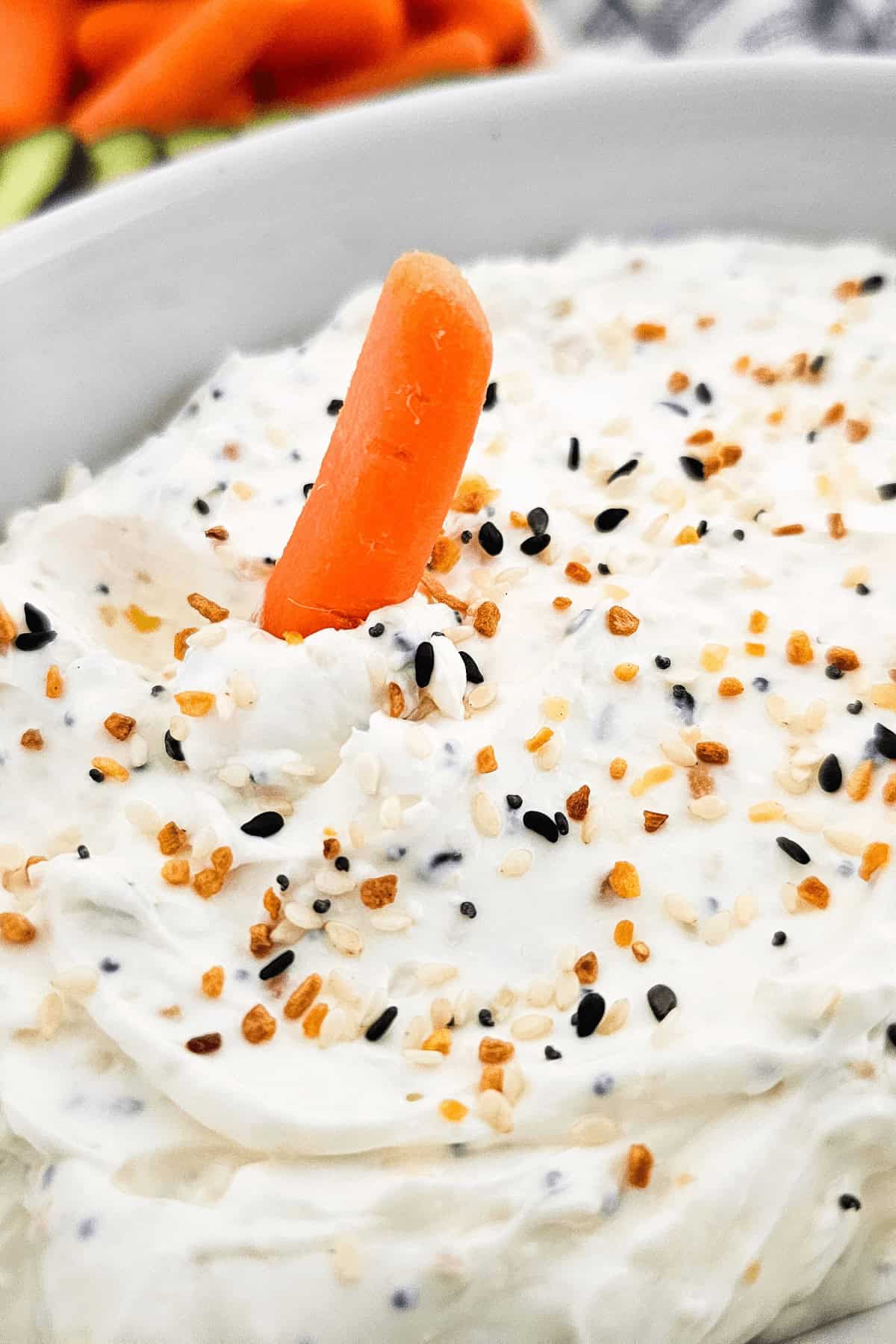 A baby carrot stands upright in a bowl of everything bagel dip, garnished with a mix of black and white sesame seeds. Blurred carrots are in the background.