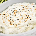 A white dish filled with creamy everything bagel dip topped with seasoning, with sliced cucumbers and baby carrots in the background.