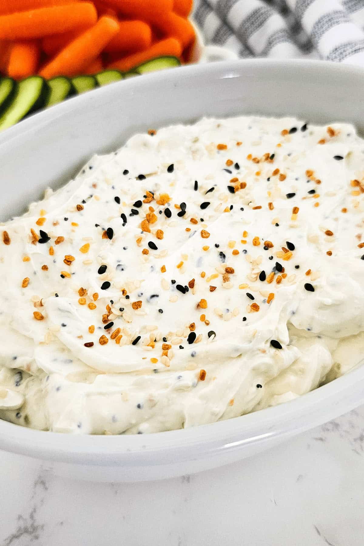 A white dish filled with creamy everything bagel dip topped with seasoning, with sliced cucumbers and baby carrots in the background.