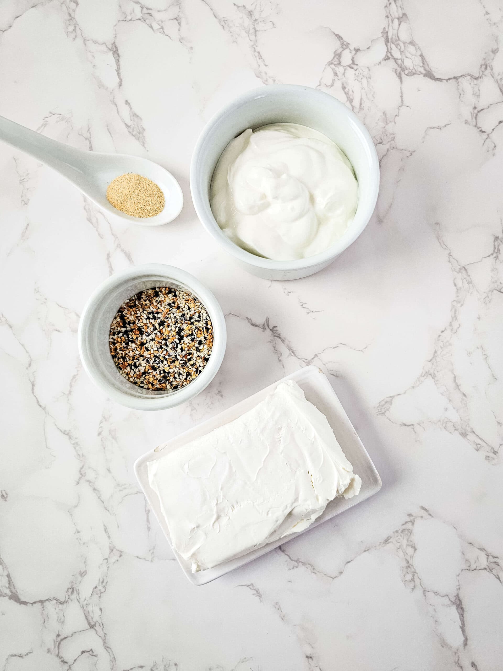 A block of cream cheese, a bowl of yogurt, a bowl of mixed spices, and a spoonful of garlic powder are arranged on a marble surface, perfect for crafting an everything bagel dip.