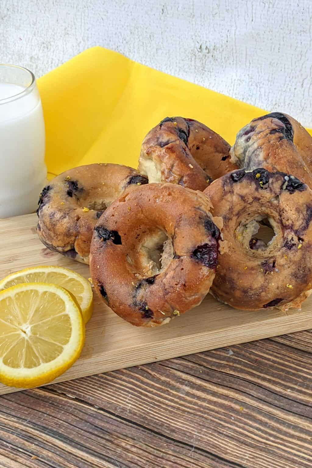 A stack of blueberry bagels on a wooden board with lemon slices and a glass of milk, set on a yellow cloth, creates a delightful scene reminiscent of freshly baked Blueberry Donuts from your favorite bakery.