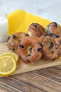 A stack of blueberry bagels on a wooden board with lemon slices and a glass of milk, set on a yellow cloth, creates a delightful scene reminiscent of freshly baked Blueberry Donuts from your favorite bakery.