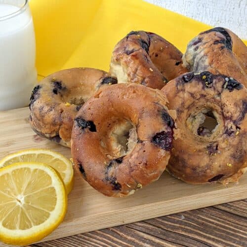 A stack of blueberry bagels on a wooden board with lemon slices and a glass of milk, set on a yellow cloth, creates a delightful scene reminiscent of freshly baked Blueberry Donuts from your favorite bakery.