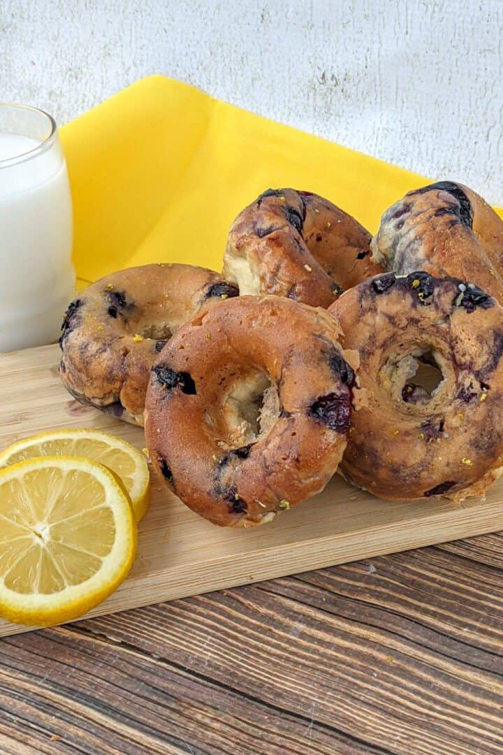 A stack of blueberry bagels on a wooden board with lemon slices and a glass of milk, set on a yellow cloth, creates a delightful scene reminiscent of freshly baked Blueberry Donuts from your favorite bakery.
