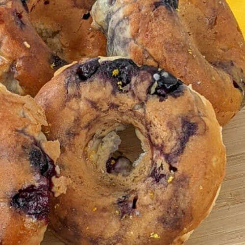 Five blueberry bagels rest on a wooden cutting board with a yellow cloth in the background, reminiscent of freshly baked blueberry donuts.