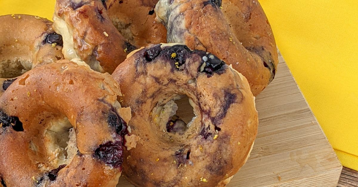 Five blueberry bagels rest on a wooden cutting board with a yellow cloth in the background, reminiscent of freshly baked blueberry donuts.
