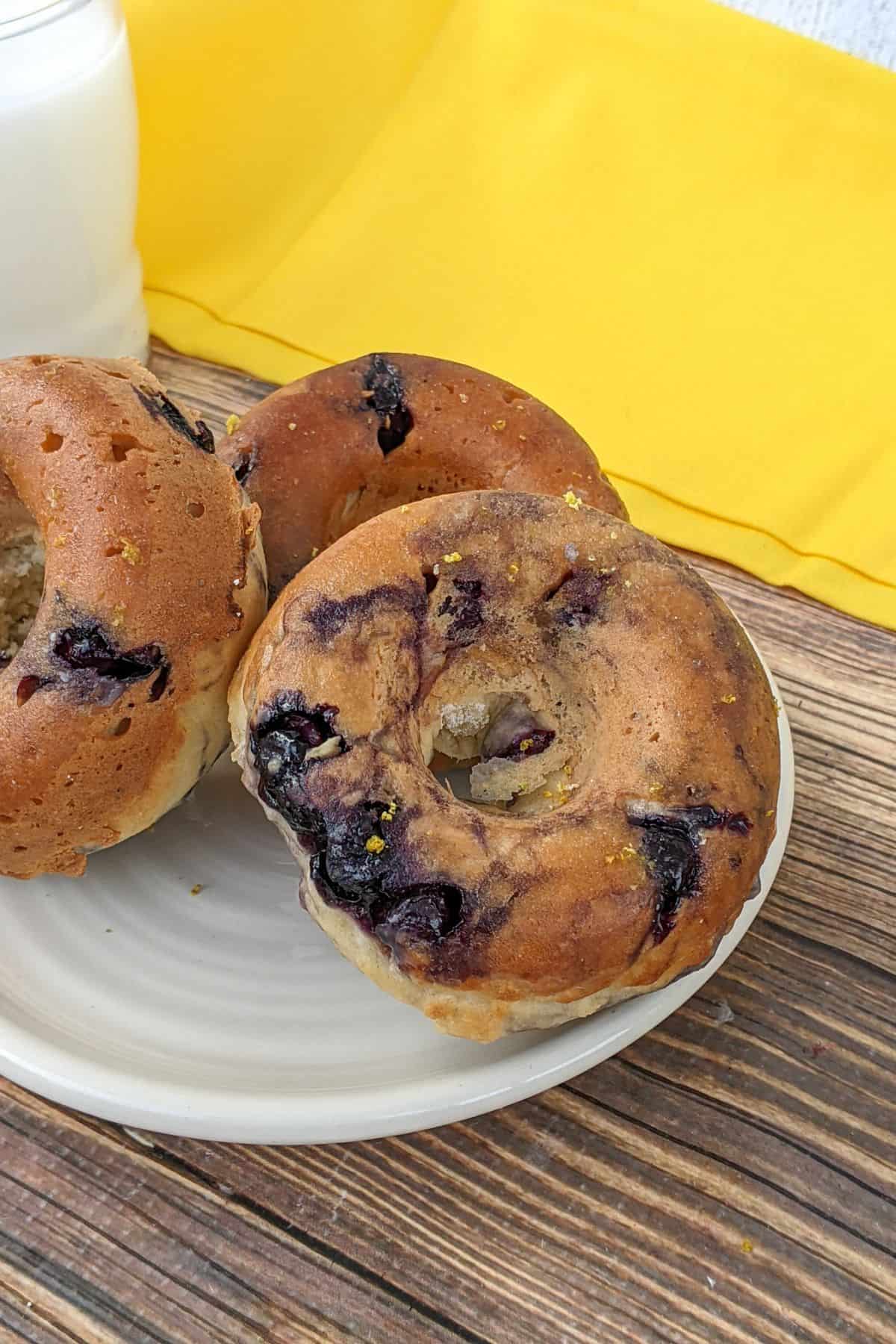 Three blueberry bagels on a white plate, set on a wooden surface with a yellow cloth and a glass of milk in the background, create an inviting scene reminiscent of fresh Blueberry Donuts from your favorite bakery.
