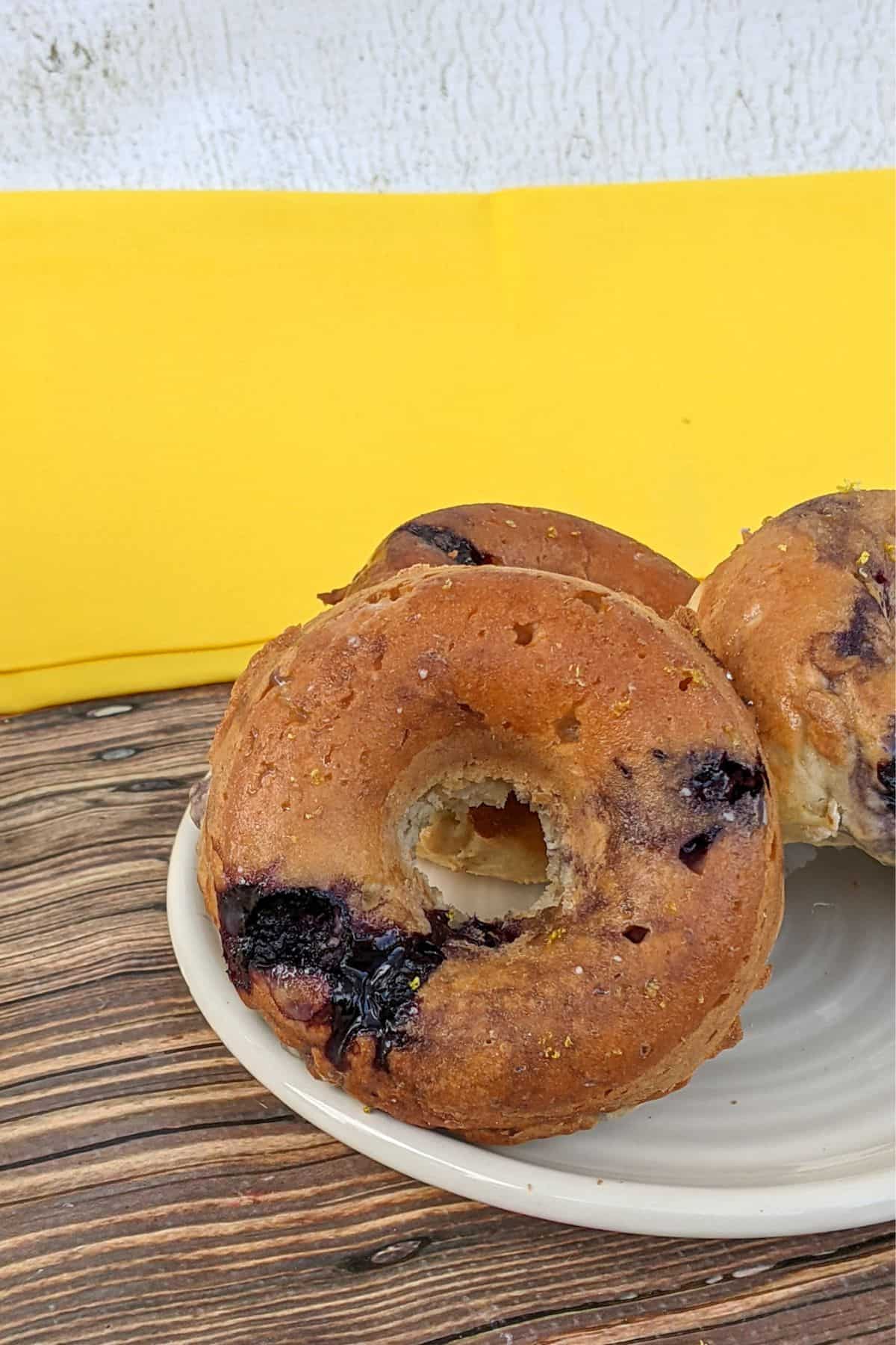 Three blueberry bagels on a white plate, set on a wooden surface with a bright yellow cloth in the background, create a scene reminiscent of freshly baked blueberry donuts enjoyed on a sunny morning.