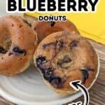 Three homemade Blueberry Donuts with a sweet blueberry glaze sit on a plate, accompanied by a glass of milk and a yellow napkin in the background. Text on image reads “Glazed Blueberry Donuts Homemade.”.