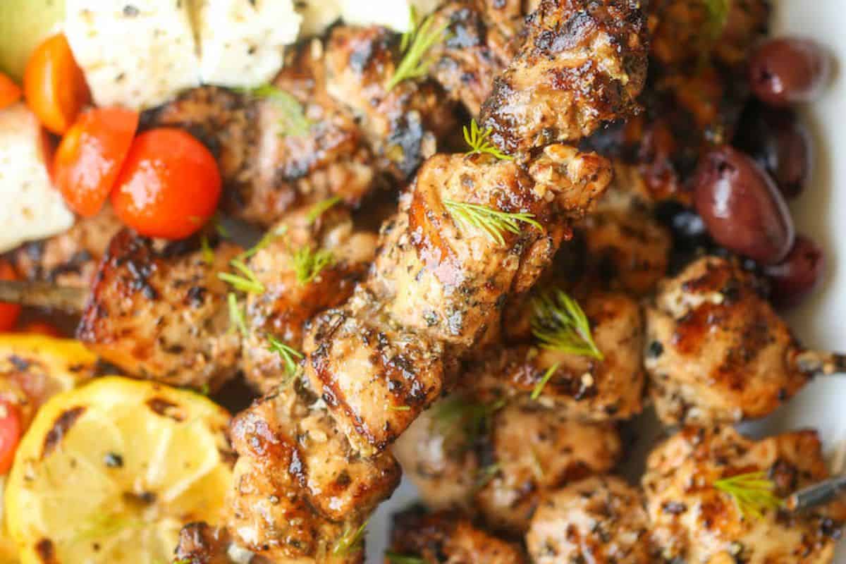 Close-up of grilled chicken kabob skewers garnished with herbs, served alongside vegetables, lemon slices, and black olives.