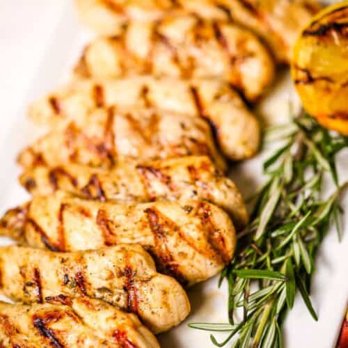 Lemon rosemary chicken tenders with perfect grill marks are arranged on a white plate, garnished with fresh rosemary and a charred lemon half.