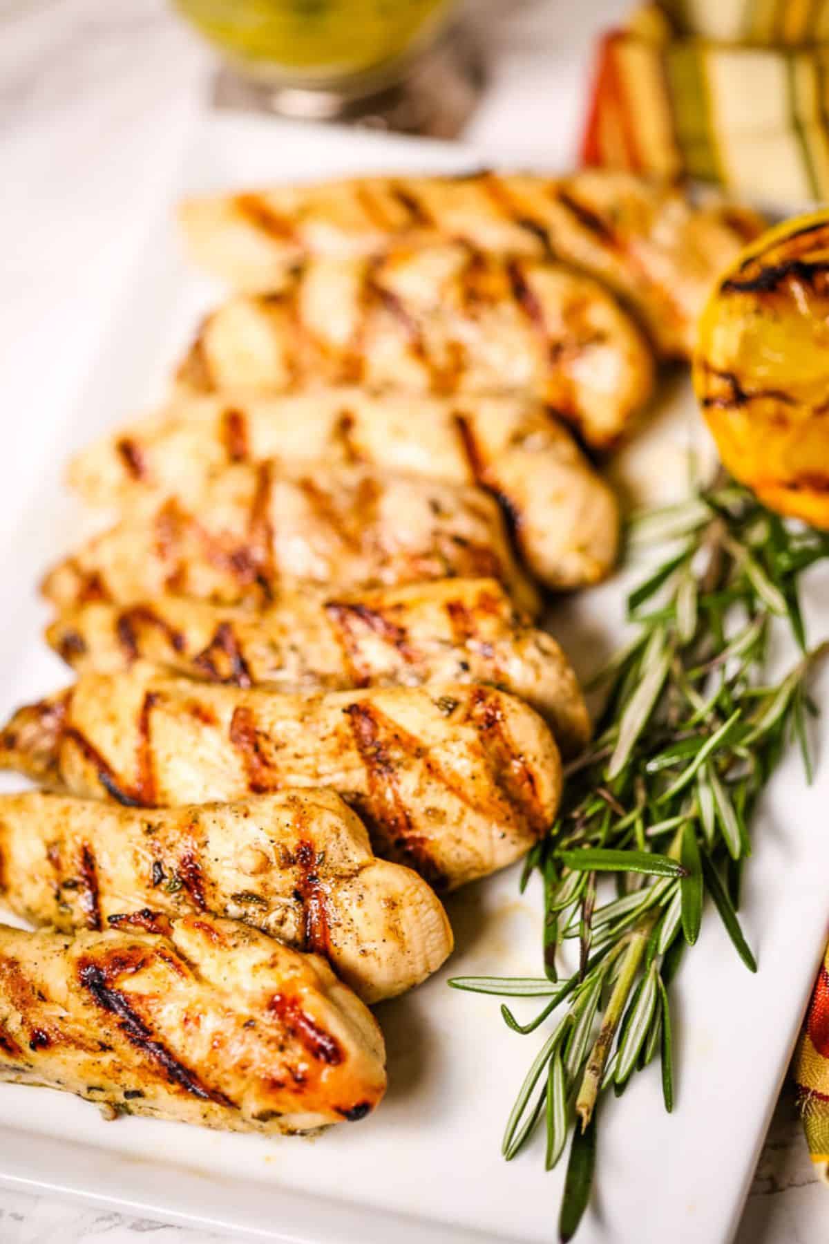 Lemon rosemary chicken tenders with perfect grill marks are arranged on a white plate, garnished with fresh rosemary and a charred lemon half.