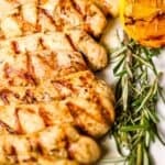 Lemon rosemary chicken breasts are arranged on a white platter with fresh rosemary and a grilled lemon half, next to a striped cloth napkin and a serving fork.