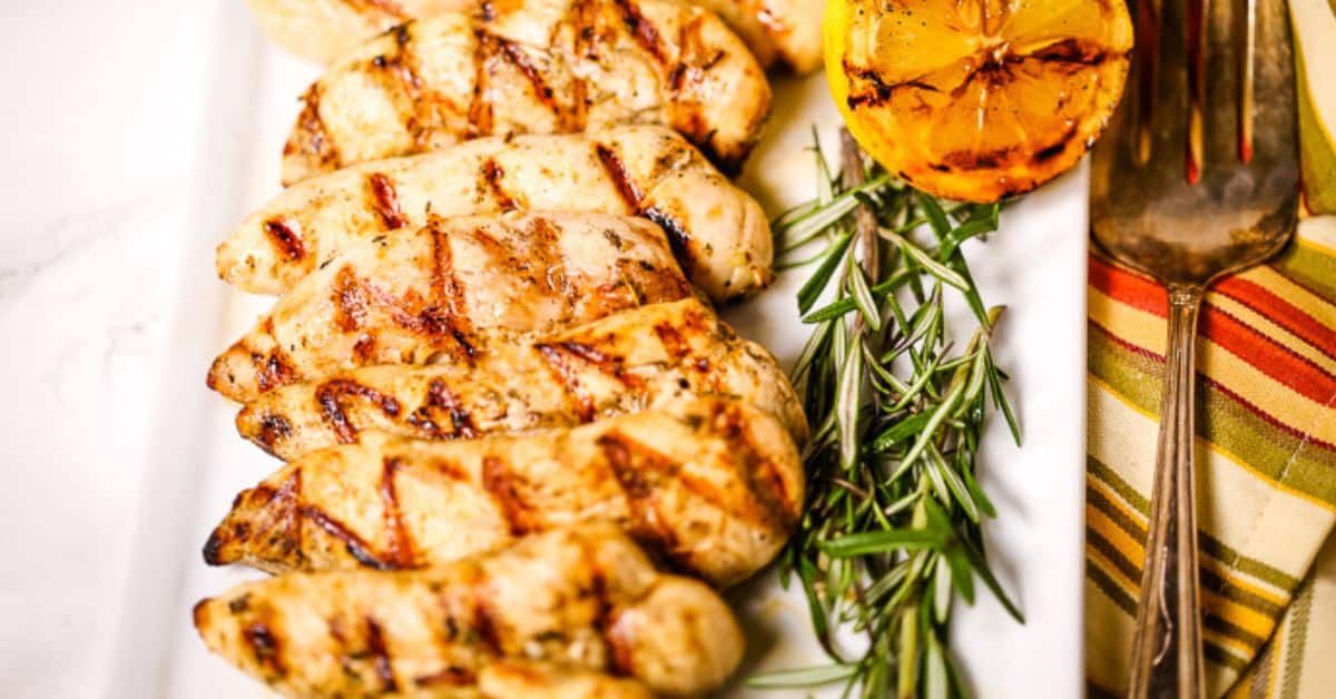 Lemon rosemary chicken breasts are arranged on a white platter with fresh rosemary and a grilled lemon half, next to a striped cloth napkin and a serving fork.