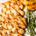 Breasts are arranged on a plate with fresh rosemary, grilled lemon, and sliced vegetables in the background.