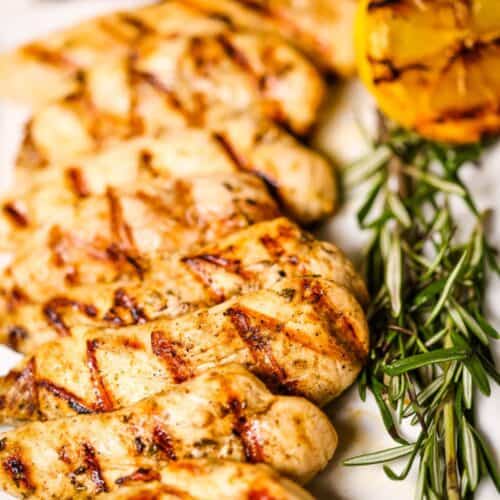 Breasts are arranged on a plate with fresh rosemary, grilled lemon, and sliced vegetables in the background.